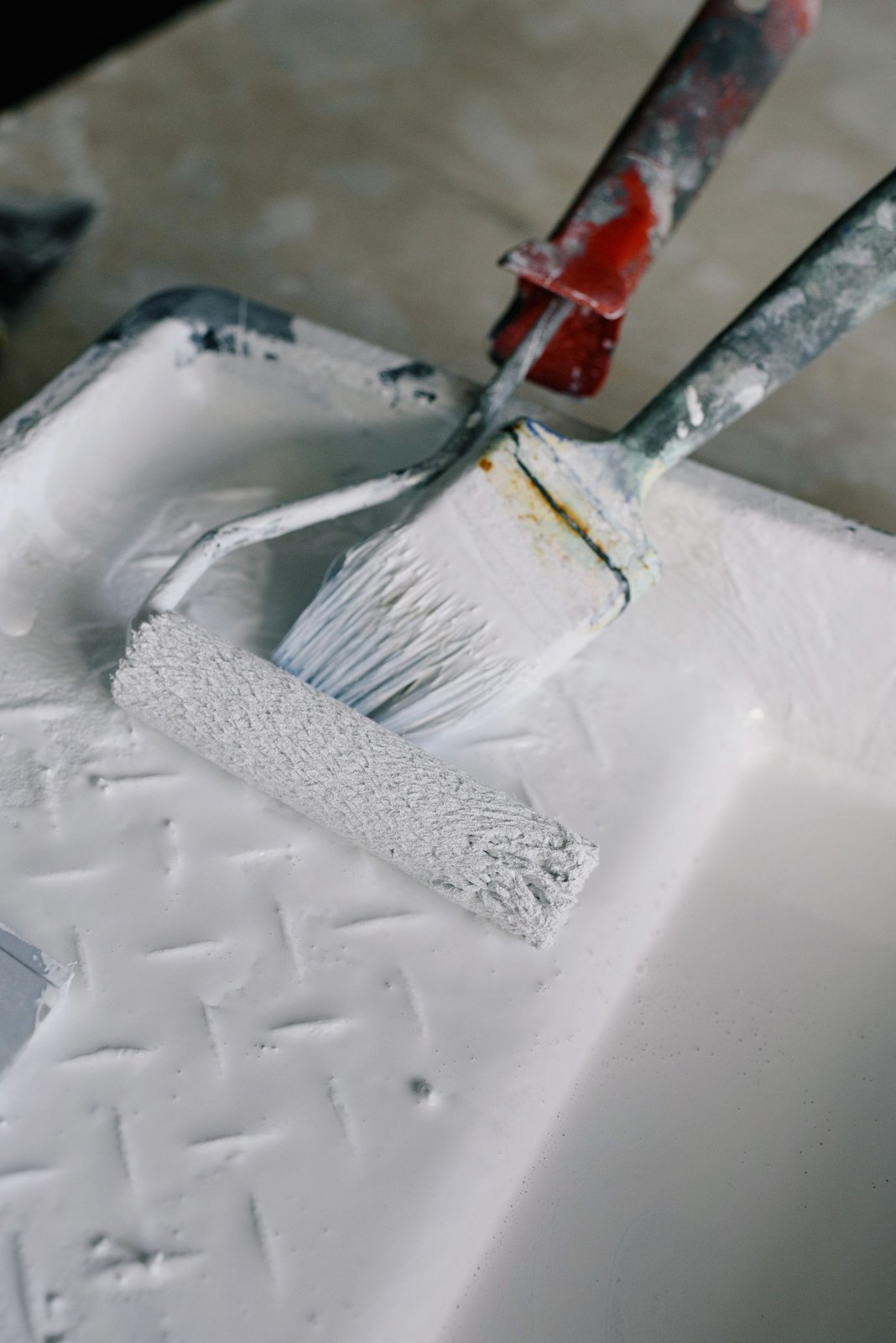 A roller and brush in a tray of white paint.